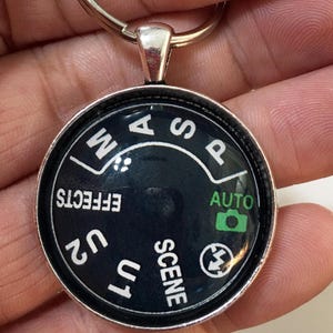 May include: A circular silver-toned keychain featuring a black camera mode dial insert. The dial displays white text including M, A, S, P, EFFECTS, U2, U1, SCENE, and a flash icon, with the word AUTO and a green camera icon printed on the right side.