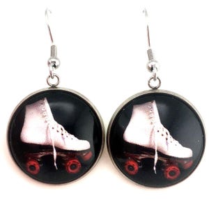 May include: A pair of round drop earrings featuring a silver-toned metal frame and hook. The center of each earring displays a circular graphic of a white roller skate with red wheels set against a solid black background, encased in a clear dome.