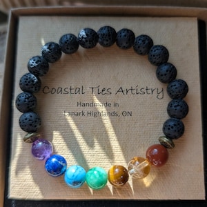 May include: A bracelet with black lava rock beads and a selection of colourful gemstone beads. The bracelet includes amethyst, blue, turquoise, green, tiger's eye, and clear quartz beads. The text "Coastal Ties Artistry" is visible.