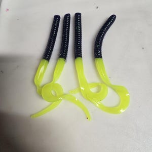 May include: Four soft plastic fishing lures. The lures have a segmented, dark blue body that transitions to a bright yellow tail. The tails are curled and flexible, designed to attract fish. The lures are designed for freshwater fishing.