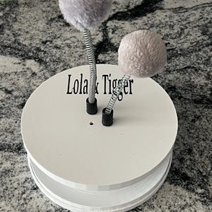May include: A white circular base with the text Lola & Tiger printed in black. Two silver metal springs are attached to the top, each topped with a soft, fuzzy, light gray pom-pom. The base features a small circular hole near the springs.