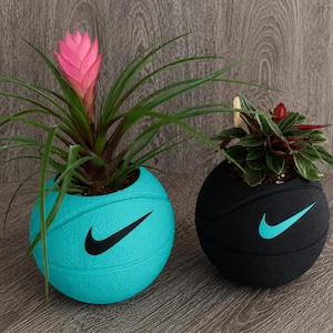 May include: Two spherical planters shaped like basketballs. The left planter is turquoise with a black Nike swoosh logo and contains a green plant with a pink flower. The right planter is black with a turquoise Nike swoosh and a green plant.