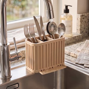 May include: A beige, rectangular, ribbed plastic cutlery drainer with three internal compartments. It features a small drainage spout at the base and is filled with stainless steel forks, knives, and spoons, positioned near a kitchen faucet.