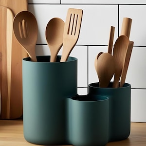 May include: A teal utensil holder with three compartments, filled with wooden spoons and spatulas. The holder sits on a wooden surface, with a white tiled background. Kitchen storage for cooking tools.