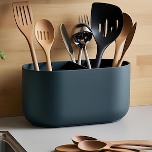 Puede incluir: Un soporte para utensilios de color verde oscuro con utensilios de cocina de madera y negros. Los utensilios incluyen espátulas, cucharas, un cuchillo y un cucharón. Dos cucharas de madera descansan sobre una tabla de madera en primer plano.