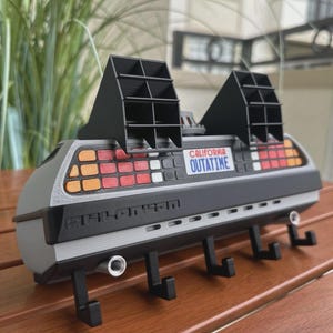 May include: A gray and black DeLorean-themed key holder with the text "CALIFORNIA OUTATIME." The key holder has four black hooks and two black vents on top. The key holder is sitting on a wooden surface.