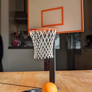 May include: A miniature basketball hoop set with an orange ball. The backboard is white with an orange border and a rectangular cutout. The net is white, and the stand is black. The ball is approximately 3.8 cm in diameter.