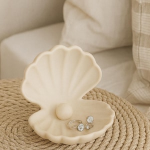 May include: A cream-colored seashell-shaped jewelry dish with a pearl and two rings with clear stones. The dish sits on a woven mat, creating a coastal theme. The rings are silver-toned.
