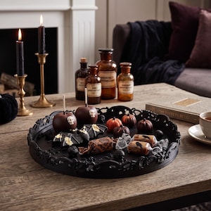 May include: A round black decorative tray with an ornate filigree border holds two chocolate-covered apples with skull accents, small orange and brown pumpkins, wrapped sweets with raven prints, and a black bat figure with gold leaf detailing.