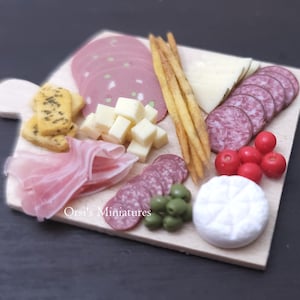May include: Miniature charcuterie board with salami, cheese cubes, prosciutto, olives, breadsticks, and red berries. The board is made of light-colored wood and is arranged on a dark surface. The text "Orsi's Miniatures" is visible.