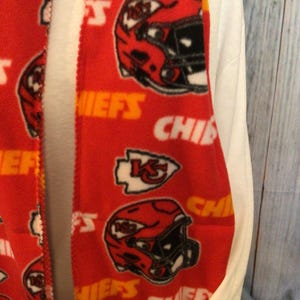 May include: Red fleece scarf with a repeating pattern of Kansas City Chiefs logos, including helmets and the team's logo. The scarf is trimmed with a yellow border.