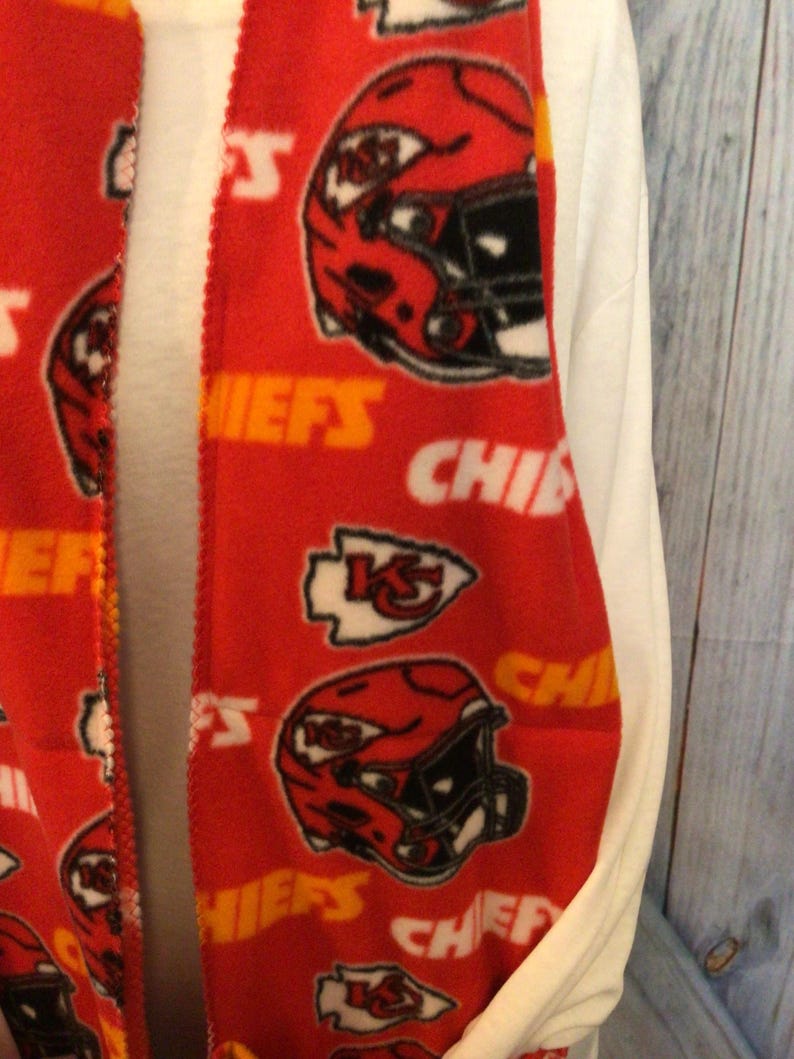 May include: Red fleece scarf with a repeating pattern of Kansas City Chiefs logos, including helmets and the team's logo. The scarf is trimmed with a yellow border.