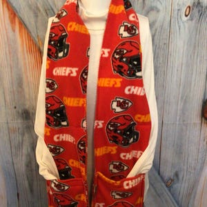 May include: Red fleece scarf with a pocket and a repeating pattern of the Kansas City Chiefs logo and the word "Chiefs".