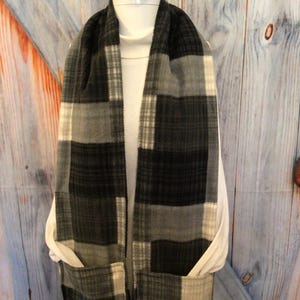 May include: A black, gray, and white plaid scarf with two pockets. The scarf is draped over a white turtleneck. The plaid pattern features large, rectangular blocks of color. The scarf is made of a soft, fleece-like material.