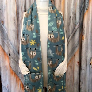 May include: Teal fleece scarf with a repeating pattern of sloths hanging from branches. The scarf has two pockets and is displayed on a mannequin. Yellow star and leaf accents complete the design.
