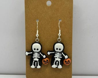 Puede incluir: Un par de pendientes de Halloween. Cada pendiente presenta una pequeña figura de esqueleto negro con detalles óseos blancos, sosteniendo una calabaza naranja. Los colgantes de esqueleto cuelgan de ganchos plateados. Presentados en una tarjeta marrón.