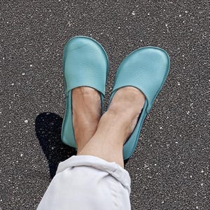 Op de afbeelding: Een paar turquoise leren instappers. De schoenen hebben een lichtblauwgroene kleur en een ronde neus. De schoenen worden getoond op een donkere, getextureerde ondergrond.