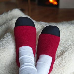 May include: A pair of red and black slippers with white socks. The slippers are made of a soft material and have a black toe cap. The socks are white and ribbed. The scene is cozy, with a fireplace in the background.