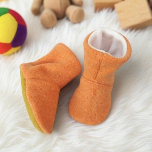 Op de afbeelding: Een paar oranje babyslofjes met een zachte, fuzzy textuur. De binnenkant is wit en de zolen zijn geelgroen. Deze slofjes zijn ontworpen om de voetjes van een baby warm en comfortabel te houden.