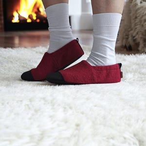 Könnte beinhalten: Rote Filzschuhe mit schwarzen Zehen, getragen mit weißen Socken auf einem flauschigen weißen Teppich. Im Hintergrund ist ein Kamin mit brennendem Feuer zu sehen. Die Schuhe sind für den Innenbereich bestimmt.