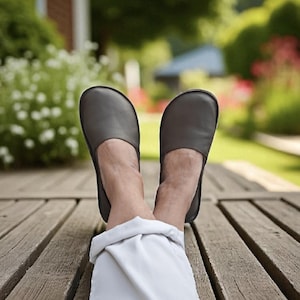 May include: A pair of gray leather slippers with a rounded toe. The slippers are worn by a person relaxing on a wooden deck. The background features greenery and a house.