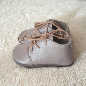 Può includere: Un paio di scarpine color talpa con lacci color cuoio. Le scarpe hanno una punta arrotondata e una morbida texture in pelle. I lacci sono annodati con un semplice nodo. Scarpe per neonati.