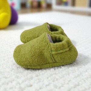 Può includere: Un paio di morbidi stivaletti verdi per bambini realizzati in un materiale strutturato. Gli stivaletti sono appoggiati su un tappeto bianco e soffice. Gli stivaletti sono progettati per mantenere i piedi del bambino caldi e comodi.