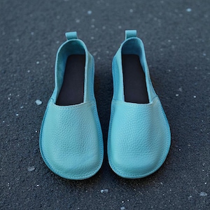 Puede incluir: Un par de zapatos sin cordones de cuero turquesa. Los zapatos tienen una punta redondeada y una superficie texturizada. El interior es marrón oscuro. Calzado casual.