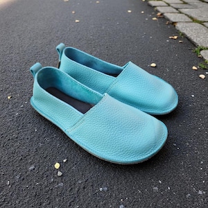 Puede incluir: Un par de zapatos sin cordones de cuero turquesa. Los zapatos tienen una puntera redondeada, una superficie texturizada y una pequeña lengüeta en el talón. Las suelas son de un tono azul más oscuro. Los zapatos miden unos 25 cm de largo.