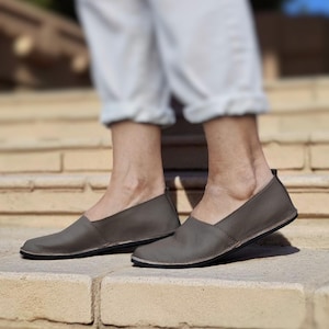 Puede incluir: Un par de zapatos grises de cuero sin cordones. Los zapatos tienen una punta redondeada y un perfil bajo, con una suela fina negra. Los zapatos se usan en los pies de una persona, con pantalones de color claro visibles.