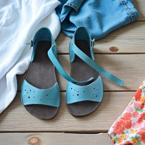 Könnte beinhalten: Ein Paar hellblaue Ledersandalen mit Riemen und Schnallenverschluss. Die Sandalen haben ein perforiertes Design auf der Vorderseite und eine dunkelbraune Sohle. Sie werden auf einer Holzoberfläche mit weißem Stoff, Denim und floralem Stoff präsentiert.