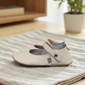 Puede incluir: Un par de zapatos de bebé de cuero color crema con correa y hebilla. Los zapatos tienen una puntera redondeada y una pequeña etiqueta que dice "DDA studio". Están colocados sobre una superficie a rayas grises y blancas.