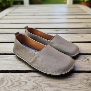 Op de afbeelding: Een paar taupekleurige instappers met een textuur van leer. De schoenen hebben een ronde neus en een laag profiel, geschikt voor casual wear. De zolen zijn donker en de binnenvoering is lichter van kleur.