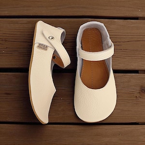 Puede incluir: Un par de zapatos Mary Jane de cuero blanco roto con correa y hebilla. Los zapatos tienen una puntera redondeada y una suela marrón. La marca "DDAA studio" es visible en un zapato. Se exhiben sobre una superficie de madera. Estilo clásico.