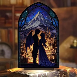 May include: A stained glass-style night light featuring a romantic scene. The image depicts a couple silhouetted against a mountain and city backdrop, with a starry sky. The light has a wooden base and a dark frame, creating a warm, inviting glow.
