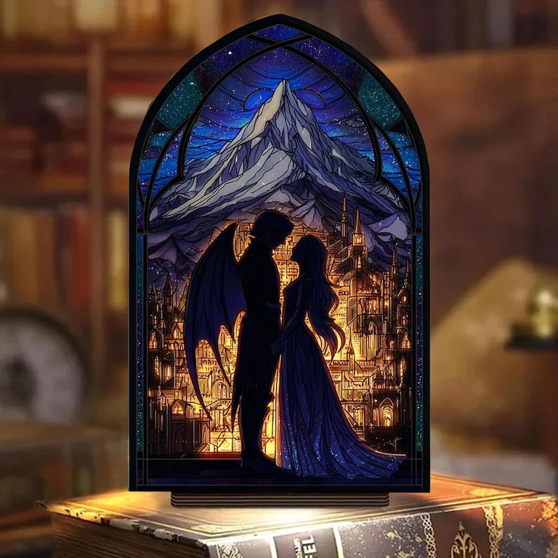 May include: A stained glass-style night light featuring a romantic scene. The image depicts a couple silhouetted against a mountain and city backdrop, with a starry sky. The light has a wooden base and a dark frame, creating a warm, inviting glow.