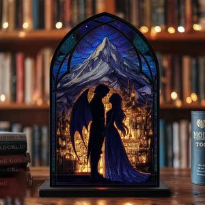 May include: A stained glass-style night light featuring a romantic scene with a winged figure and a woman in front of a mountain and city backdrop. The artwork is framed in a gothic arch shape, with a dark wooden base.