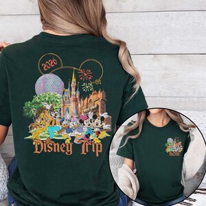May include: Dark green t-shirt featuring a colorful Disney Trip design with Mickey and Minnie Mouse, Donald and Daisy Duck, Goofy, and Pluto. The design includes the text "Disney Trip" and the year "2026". A smaller version of the design is on the front.