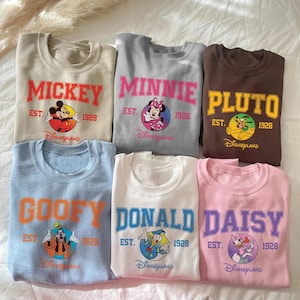 May include: A collection of six crewneck sweatshirts in various colors, each featuring a different Disney character's name in bold lettering. The sweatshirts include Mickey, Minnie, Pluto, Goofy, Donald, and Daisy, with the Disneyland logo and the year 1928.