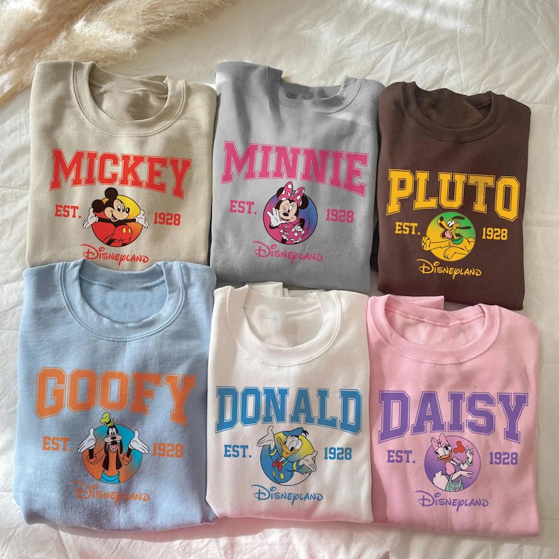 May include: A collection of six crewneck sweatshirts in various colors, each featuring a different Disney character's name in bold lettering. The sweatshirts include Mickey, Minnie, Pluto, Goofy, Donald, and Daisy, with the Disneyland logo and the year 1928.