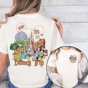 May include: Cream-colored t-shirt featuring a colorful Disney Trip graphic on the back, including Mickey and Minnie Mouse, Donald Duck, and Goofy. The design incorporates the year 2026, a castle, and the text "Disney Trip."