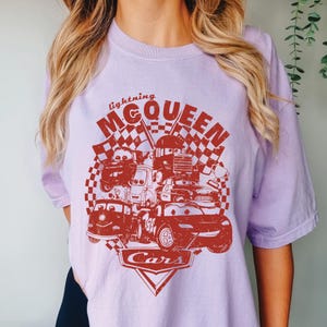 May include: A light purple t-shirt featuring a red graphic of characters from the movie Cars, including Lightning McQueen. The design includes the text "lightning McQueen" and the word "Cars" in a triangle.