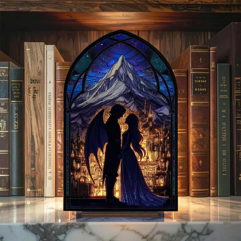 May include: A stained glass panel depicting a couple in silhouette with a mountain and city backdrop. The panel features a dark blue night sky with stars and a mountain range. The couple is in front of a city with illuminated windows. The panel is set against a backdrop of books.