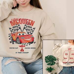 May include: Beige sweatshirt featuring a graphic of Lightning McQueen from the movie Cars, with checkered flags and text. The back of the sweatshirt displays the number 95 in red. The sweatshirt is a crew neck style.