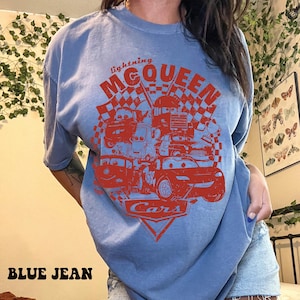 May include: Blue jean t-shirt featuring a red graphic of Lightning McQueen and other characters from the movie Cars. The graphic includes the text "Lightning McQueen" and "Cars". The shirt is a light blue color.