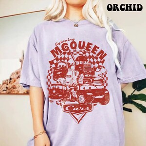 May include: A light purple t-shirt featuring a red graphic of Lightning McQueen and other characters from the movie Cars. The design includes the text "Lightning McQueen" and "Cars" with checkered flags.