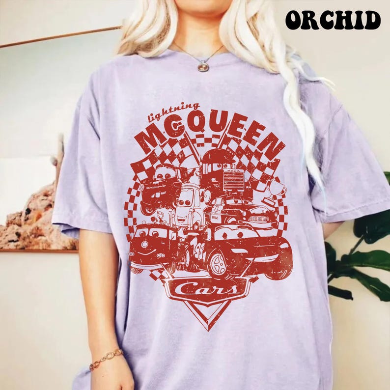 May include: A light purple t-shirt featuring a red graphic of Lightning McQueen and other characters from the movie Cars. The design includes the text "Lightning McQueen" and "Cars" with checkered flags.