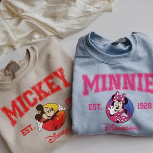 May include: Two sweatshirts, one tan and one light blue, featuring Disney characters. The tan sweatshirt has "MICKEY" in red lettering with a graphic of Mickey Mouse. The blue sweatshirt says "MINNIE" in pink, with Minnie Mouse. Both say "EST. 1928" and "Disneyland".