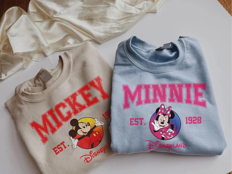 May include: Two sweatshirts, one tan and one light blue, featuring Disney characters. The tan sweatshirt has "MICKEY" in red lettering with a graphic of Mickey Mouse. The blue sweatshirt says "MINNIE" in pink, with Minnie Mouse. Both say "EST. 1928" and "Disneyland".