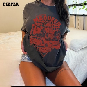 May include: Dark gray t-shirt featuring a red graphic of race cars and the text "McQueen Cars". The shirt has short sleeves and a crew neck. The word "PEEPER" is printed in the upper left corner.